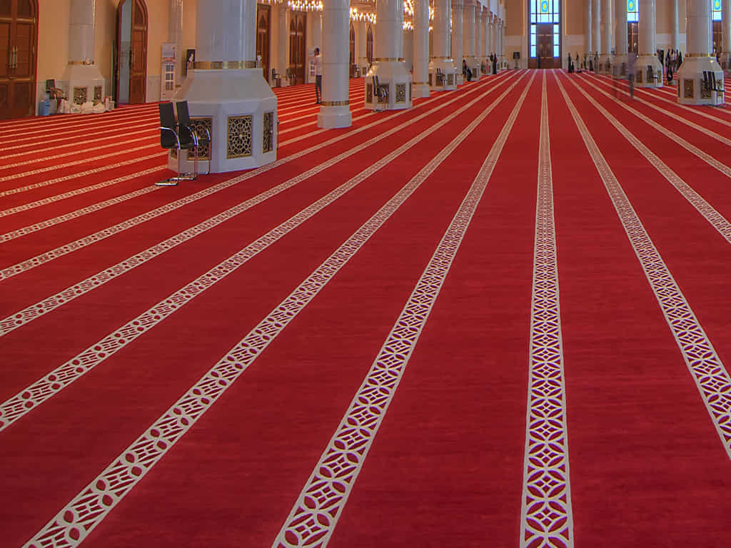 A large, red prayer carpet with geometric patterns fills a spacious mosque hall