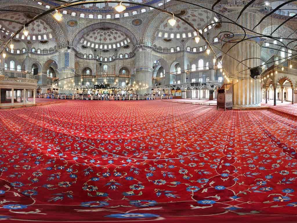 The mosque is filled with people, and the walls and ceiling are decorated with intricate blue and white tiles